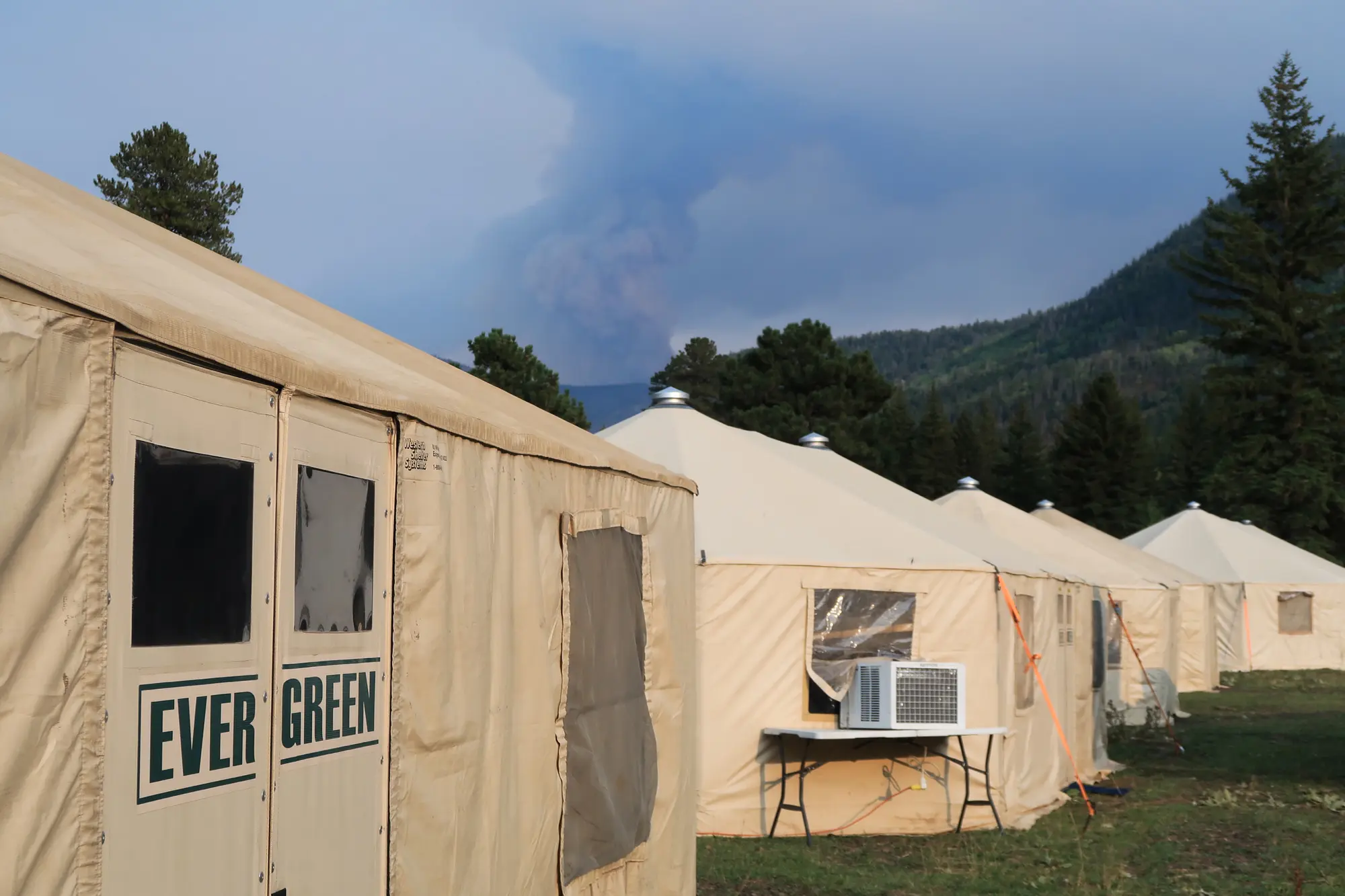 A row of Evergreen Resource yurts at a mountainside base camp, smoke rising on the ridge